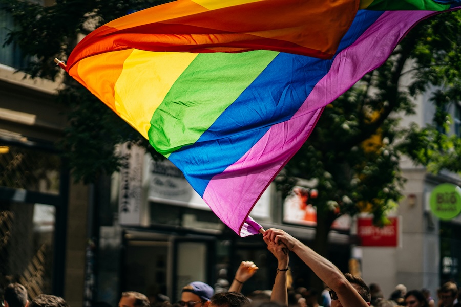 Crowd waving rainbow pride flag at an LGBTQ+ event, buildings and trees in background