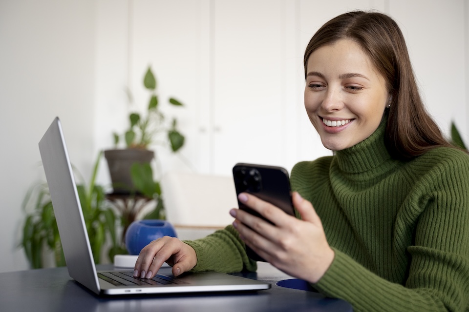 Happy woman working on laptop, reviewing her phone. Texas SMS marketing changes.
